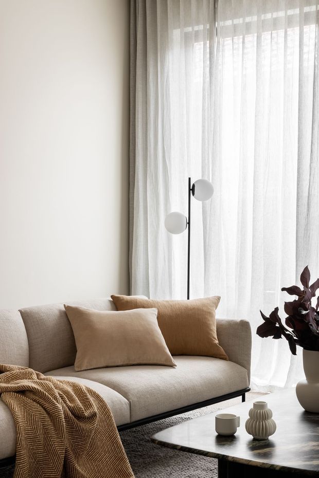 A softly lit contemporary living room featuring a neutral upholstered sofa, beige cushions, a textured brown throw, sheer full-height curtains, a slender black floor lamp, and a dark stone coffee table with ceramic vessels. A potted dark-leaved plant adds contrast beside the window.
