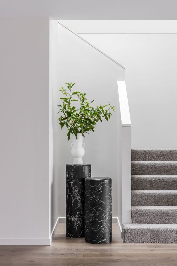 A minimalist interior stairway with a small recessed display niche beside carpeted stairs. The niche contains two tall black marble-look cylindrical plinths, one supporting a white vase with leafy branches. White walls, pale timber flooring, and grey carpet create a restrained monochromatic palette.