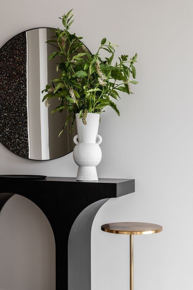A refined interior vignette featuring a sculptural black console table with large arched supports, a white ceramic urn filled with leafy greenery, an asymmetrical rounded mirror, and a slim brass-and-brown side table. The palette is monochromatic with warm metallic accents and strong geometric forms.