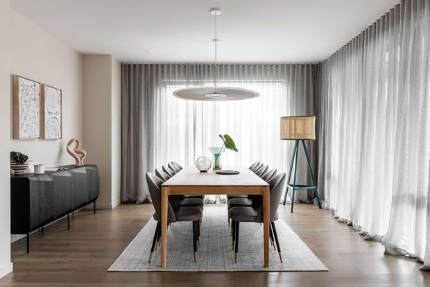 A refined dining room with a rectangular timber dining table and upholstered chairs on a large grey rug. Full-height sheer and grey curtains surround expansive glazing, while a dark sideboard, sculptural decor, pendant light, and tripod floor lamp complete the calm neutral interior.
