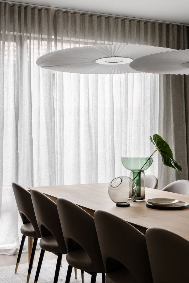 A refined dining room with a long light timber dining table surrounded by sculptural dark upholstered chairs. Full-height sheer and taupe curtains cover the windows, while a large white pleated pendant light hangs overhead. Minimal glass and ceramic table decor adds a restrained, elegant finish.