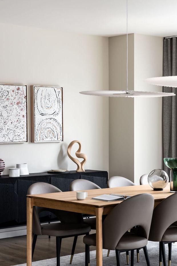 A refined contemporary dining room with a large natural timber dining table surrounded by upholstered curved-back chairs. A dark built-in sideboard lines the wall beneath two abstract artworks and sculptural decor. A slim sculptural pendant light hangs above the table, with neutral plaster walls, dark curtains, and timber flooring creating a calm, sophisticated setting.