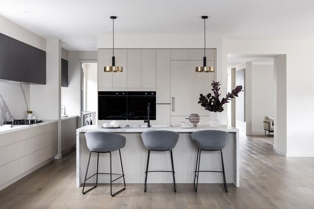 A bright, contemporary open-plan kitchen with a large pale island and three grey upholstered bar stools. The kitchen features flat-panel light cabinetry, integrated black appliances, a pale benchtop, dark wall cabinetry, pendant lighting, and light timber flooring. An open doorway leads toward a furnished adjoining living area.
