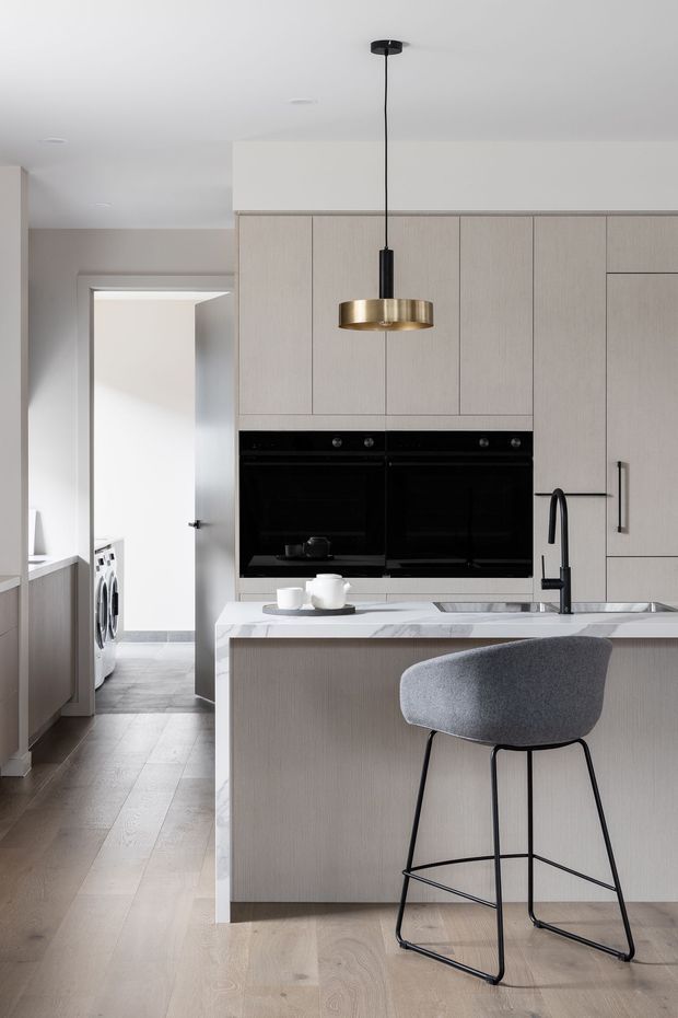 A refined contemporary kitchen with a pale timber-look island and full-height cabinetry, integrated black double ovens, a white veined stone benchtop, undermount sink and dark tapware. A brass pendant light hangs above the island, paired with a grey upholstered bar stool. Light timber flooring, soft neutral walls and a partially visible laundry area create a clean, understated interior.