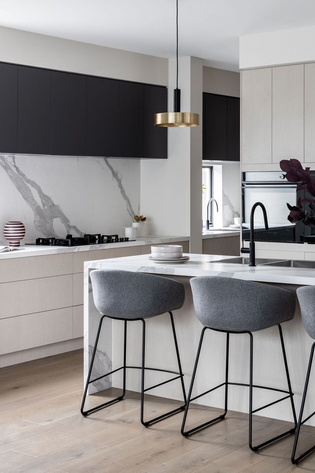 A refined contemporary kitchen with a large central island, pale timber cabinetry, white marble-look veined surfaces, and matte black overhead cabinetry. Three grey upholstered bar stools with black metal frames line the island. A brass pendant light hangs above the island, complemented by black tapware and appliances. The space has light timber flooring, integrated appliances, and a restrained neutral palette with dark accents.
