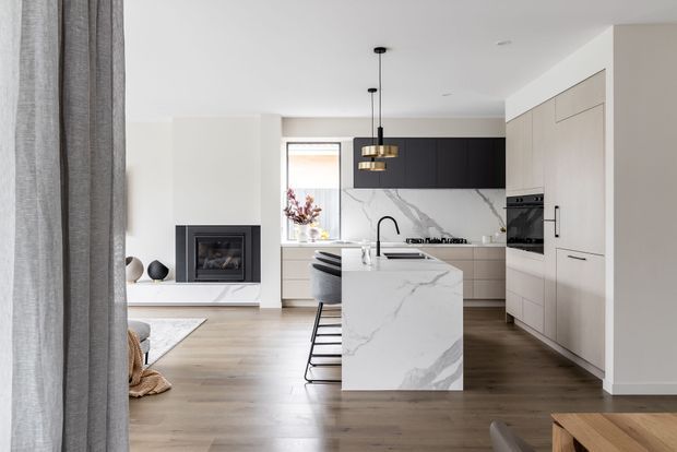 Bright contemporary open-plan kitchen and living area viewed from behind a grey curtain. The kitchen features a large white marble-look island with an integrated sink and black tap, pale cabinetry, dark overhead cupboards, built-in appliances, a marble-look splashback, pendant lighting, and timber flooring. A fireplace and seating area are visible to the left.