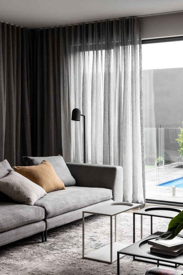 A refined contemporary living room with a large grey sectional sofa, textured neutral rug, slim black-framed nesting coffee tables, black floor lamp, and layered dark and sheer full-height curtains. Floor-to-ceiling glazing opens to an outdoor area with a visible swimming pool. The room has a restrained neutral palette with a warm tan cushion and a green plant accent.