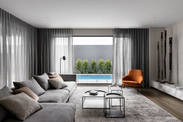 A refined contemporary living room with a large grey sectional sofa, layered neutral cushions, slim black-framed coffee tables, an orange accent lounge chair, full-height sheer curtains, and a low white media console. Floor-to-ceiling glazing opens onto a pool terrace with greenery beyond, while tall sculptural objects decorate the wall beside the television.
