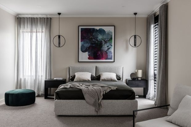 A refined contemporary bedroom with a centered upholstered bed, grey textured headboard and matching bed base, dark bedding, bedside tables, pendant lights, sheer curtains, and layered full-height window treatments. Neutral taupe, grey, black, and white tones are complemented by abstract wall art and a small deep teal ottoman.