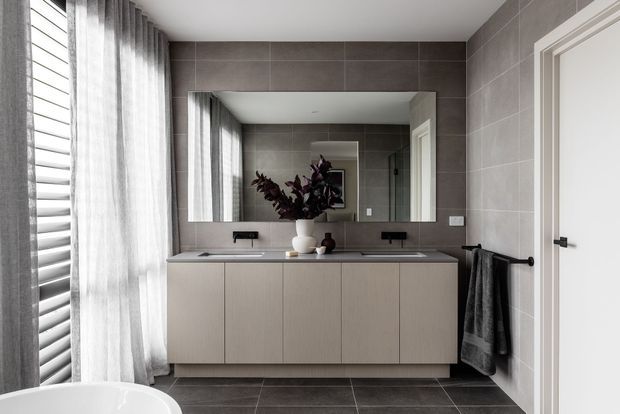 Contemporary bathroom with a wide floating double vanity, integrated basins, large frameless mirror, and dark grey tiled walls and floor. Soft daylight filters through sheer curtains and horizontal blinds beside the bathtub, while a white panel door and dark towel rail add contrast. The restrained palette and clean lines create a refined, calm atmosphere.