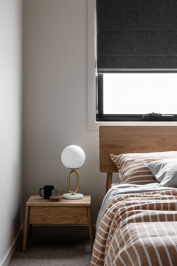 A close-up of a warmly styled bedroom corner featuring a timber bedhead, matching timber bedside table, upholstered bed with brown-and-white striped bedding, and a small round white globe lamp with a brass-coloured base. A dark roller blind covers the window above the bed, while neutral painted walls and grey carpet create a calm, contemporary feel.