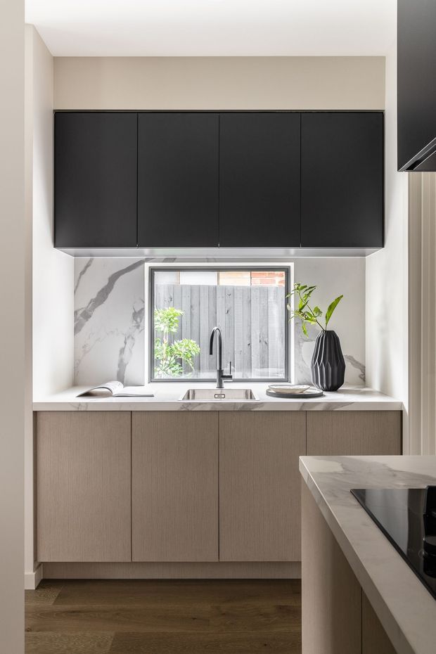 Compact contemporary kitchen with a refined two-tone scheme. Matte black upper cabinetry contrasts with warm taupe wood-grain lower cabinetry, while a white marble-look splashback and benchtops frame the sink beneath a window. A partial island with an integrated cooktop is visible in the foreground, with timber flooring and restrained decorative styling.