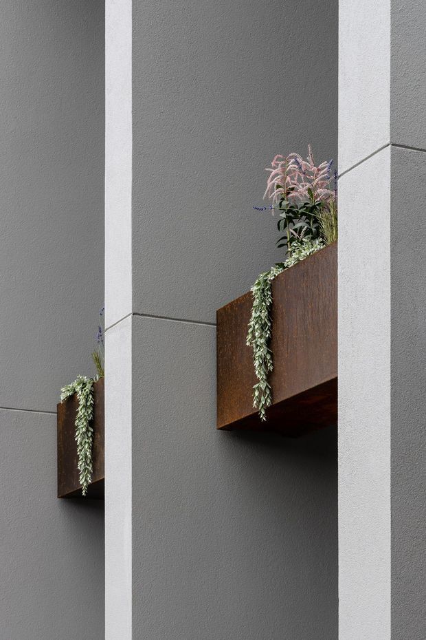 Close-up architectural detail of a contemporary grey rendered facade with strong vertical white structural bands and projecting rusted steel planter boxes containing cascading and upright greenery. The composition emphasizes minimalist geometric forms, textured wall surfaces, and integrated landscaping.