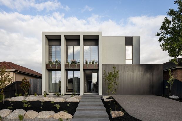 Contemporary multi-storey residential facade with a pale rendered upper volume, tall vertically proportioned glazed openings, dark metal or timber window surrounds, and a darker textured garage volume. A stepped path leads through minimalist rock landscaping to the entrance, with an adjoining driveway and neighbouring homes visible.