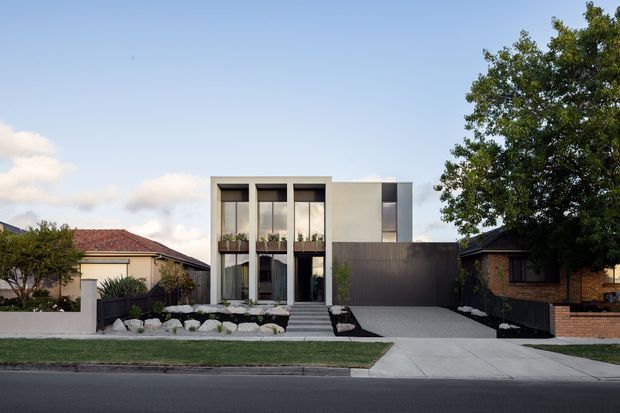 Street-facing view of a contemporary multi-level residence with a crisp white rendered facade, tall narrow windows, dark vertical-clad garage volume, and a broad concrete driveway. The modern home is set among established suburban properties, with landscaped rock beds, a lawn, fencing, and mature trees.