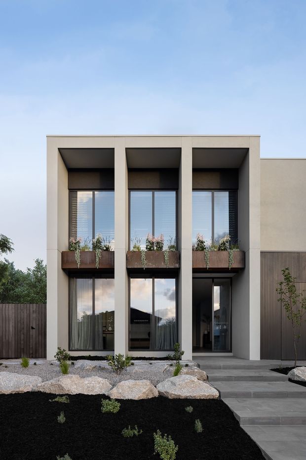 Front exterior view of a contemporary two-storey residence with a rectilinear rendered facade, three tall recessed window bays, dark-framed glazing, integrated planter boxes, and a recessed entry. The property features a minimalist landscaped frontage with dark mulch, pale gravel, boulders, timber fencing, and broad concrete steps.