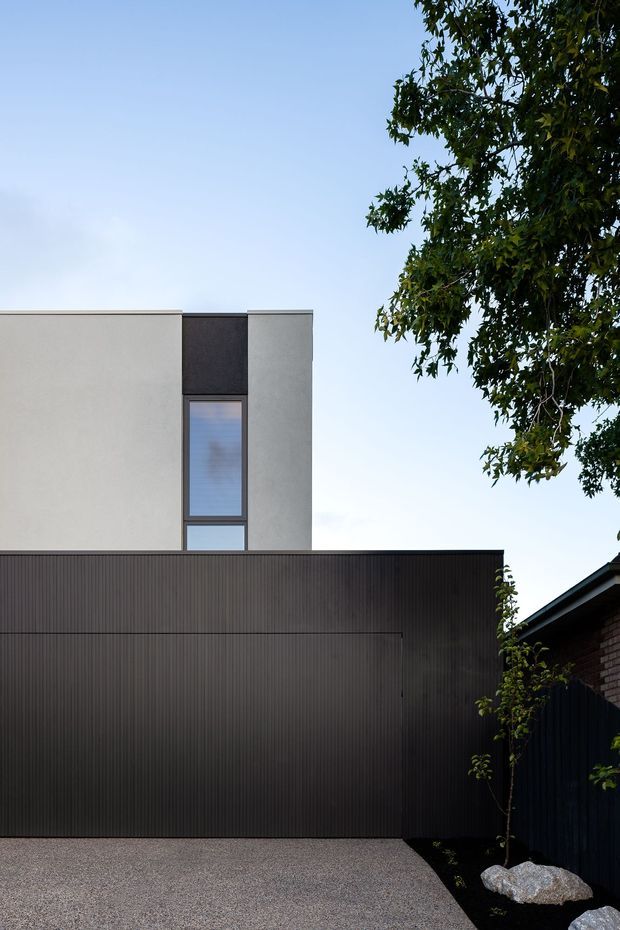 A contemporary urban residential facade viewed from the driveway. The building features a simple flat-roofed upper volume in pale grey rendered finish, a narrow vertical window opening, and a large dark vertically ribbed facade or garage door at ground level. A neighbouring brick structure, dark fence, gravel driveway, rocks, and mature tree frame the composition.