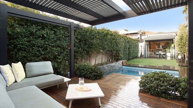 A landscaped outdoor entertaining area with a dark timber deck, pergola with slatted roof, outdoor lounge seating, raised garden beds and a narrow swimming pool. The view looks toward a glazed indoor entertaining space beyond the pool, with dense hedging providing privacy.