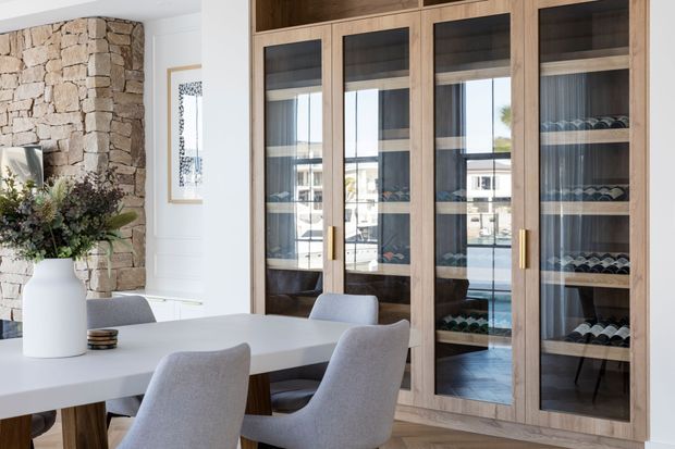 Bright, refined dining area with a large white dining table and upholstered grey chairs. A full-height timber wine display and storage wall with glass-fronted doors dominates the background, while a natural stacked-stone feature wall and white-painted walls add textural contrast. Daylight enters through the glazing and reflects across the cabinetry.
