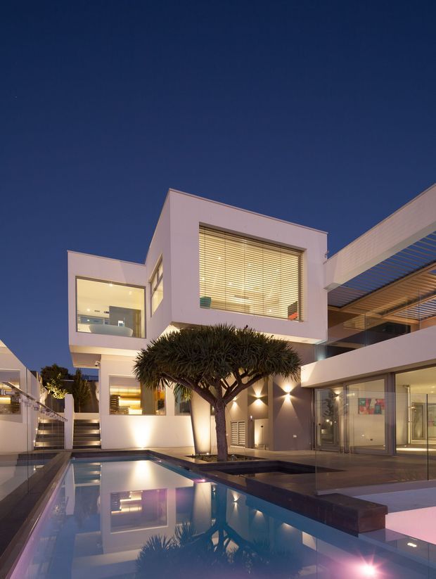 Twilight exterior view of a sophisticated multi-level modern residence arranged around a reflective swimming pool. The building features crisp white rendered cubic forms, extensive glazing, horizontal window blinds, a covered outdoor terrace with slatted canopy, recessed lighting, and a sculptural trimmed tree in the courtyard.