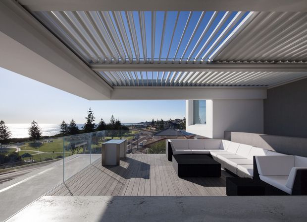A contemporary rooftop or upper-level terrace overlooking a coastal suburban landscape. The space features a broad timber deck, frameless glass balustrade, white and charcoal exterior walls, and a large outdoor sectional lounge arranged beneath a slatted pergola with a retractable or louvred roof. The setting is bright and open, with ocean visible in the distance.