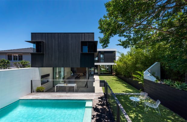Contemporary multi-level residential home with a dark vertically clad upper volume, extensive glazing, and a cantilevered form overlooking a turquoise swimming pool. The landscaped yard includes a narrow lawn, raised planting beds, outdoor dining furniture, and mature trees, while a neighbouring light-coloured wing is partially visible on the left.