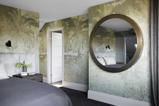 A contemporary bedroom with a large bed in the foreground, botanical wallpaper covering the walls, a white panelled door, dark carpet, and a round bronze-framed wall mirror reflecting the bed and opposite wall. Black wall lights, a small bedside table, and a potted plant complete the room.