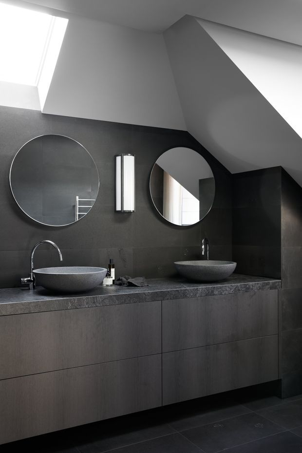 A dramatic contemporary bathroom featuring a long dark floating vanity with two round vessel basins, dark stone benchtop and wall finish, circular mirrors, slim vertical wall lighting, and a sculptural angular ceiling form with a skylight. The monochromatic charcoal and grey palette creates a refined, moody atmosphere.