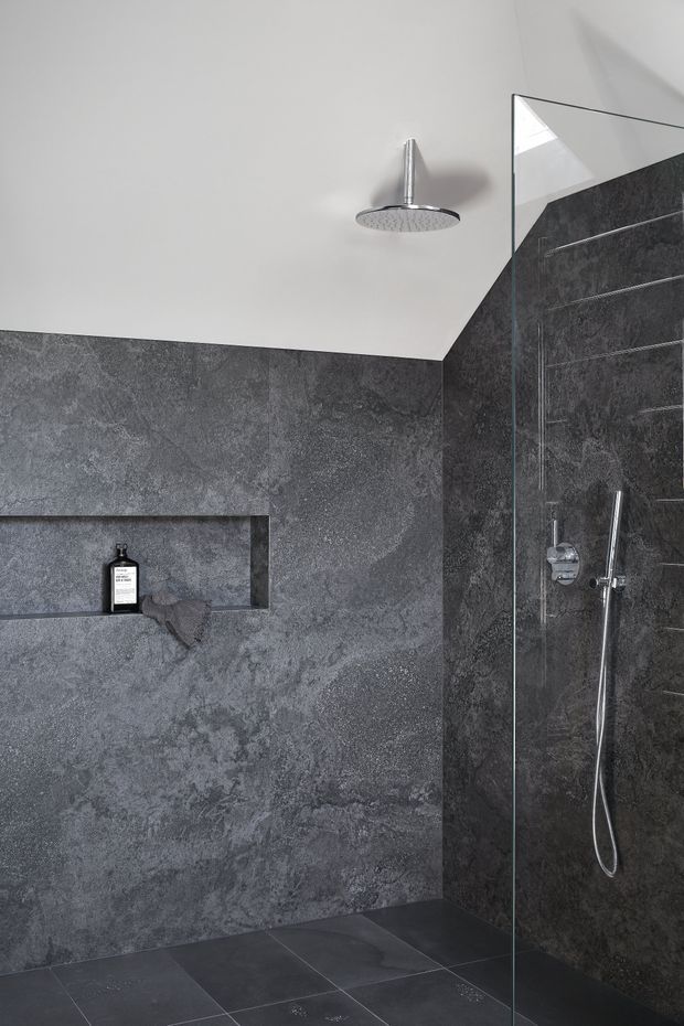 A modern walk-in shower enclosed by a frameless glass panel. The walls are clad in dark grey textured stone-look slabs with a recessed storage niche, while the floor uses large dark tiles. Chrome rain shower and handheld fittings contrast with the light painted ceiling, creating a restrained, monochromatic bathroom interior.