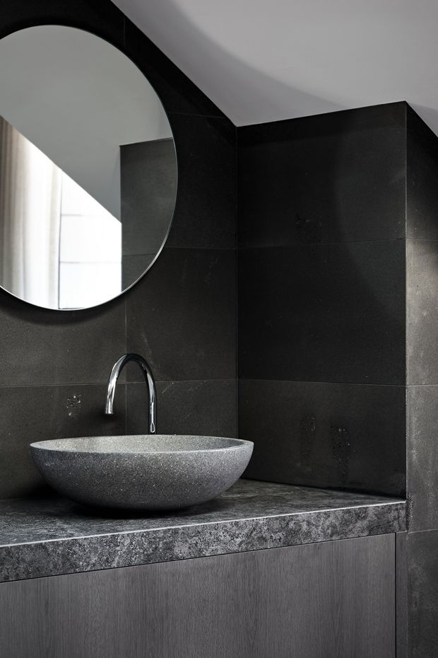 Close-up of a dark, refined bathroom vanity featuring a round vessel basin, polished chrome gooseneck tap, large round mirror, and black textured wall tiles. The basin sits on a mottled dark stone benchtop above vertically textured dark cabinetry, with soft daylight entering from the left.