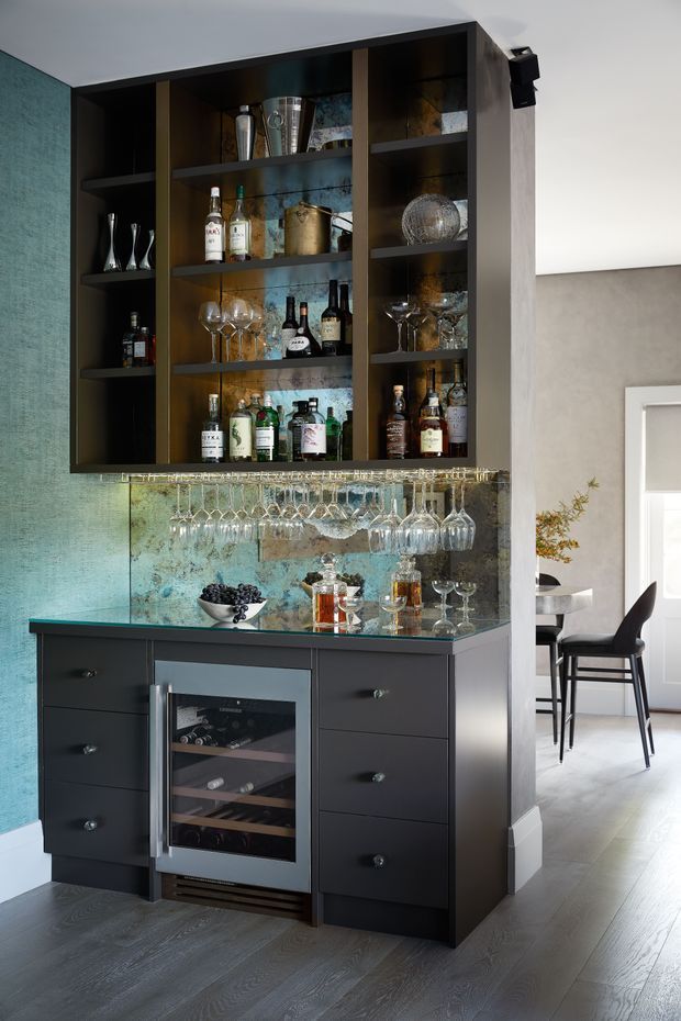 A built-in home bar occupies a tall alcove, with dark charcoal cabinetry, open shelving filled with bottles and glassware, mirrored blue-green backsplash panels, and an under-counter wine refrigerator. Stemware hangs beneath the reflective benchtop, while a dining area is visible through the opening to the right. The space has a refined contemporary look with dark joinery and cool-toned finishes.