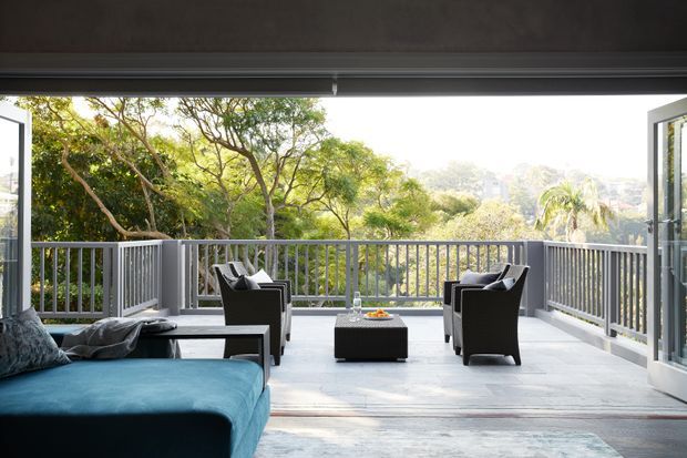 A bright, open-sided outdoor terrace or balcony viewed from an adjoining interior, furnished with a large blue chaise sectional, four dark lounge chairs, and a central coffee table. Grey tile flooring, slim grey metal balustrades, wide opening glazing, and a broad overhanging ceiling frame the leafy suburban landscape beyond.