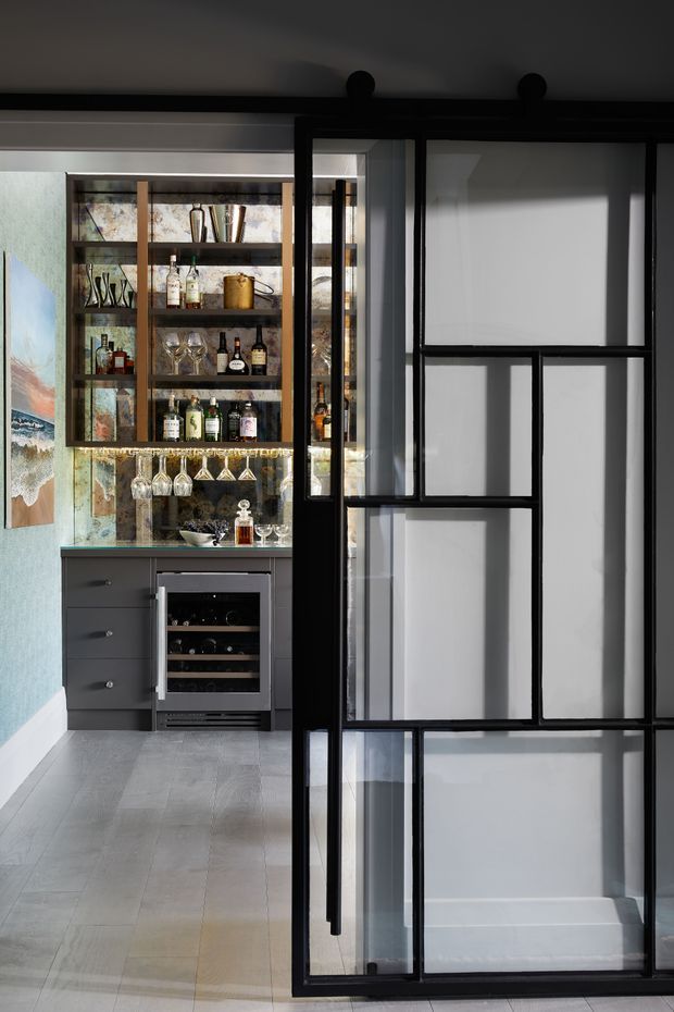 A compact, sophisticated home bar or drinks station viewed through a black metal-framed glass partition door. The space features dark built-in cabinetry, open timber shelving stocked with bottles and glassware, a wine refrigerator, a reflective patterned backsplash, pale timber-look flooring, and a muted blue wall. The strong geometric glazing and dark framing create an industrial-modern character.
