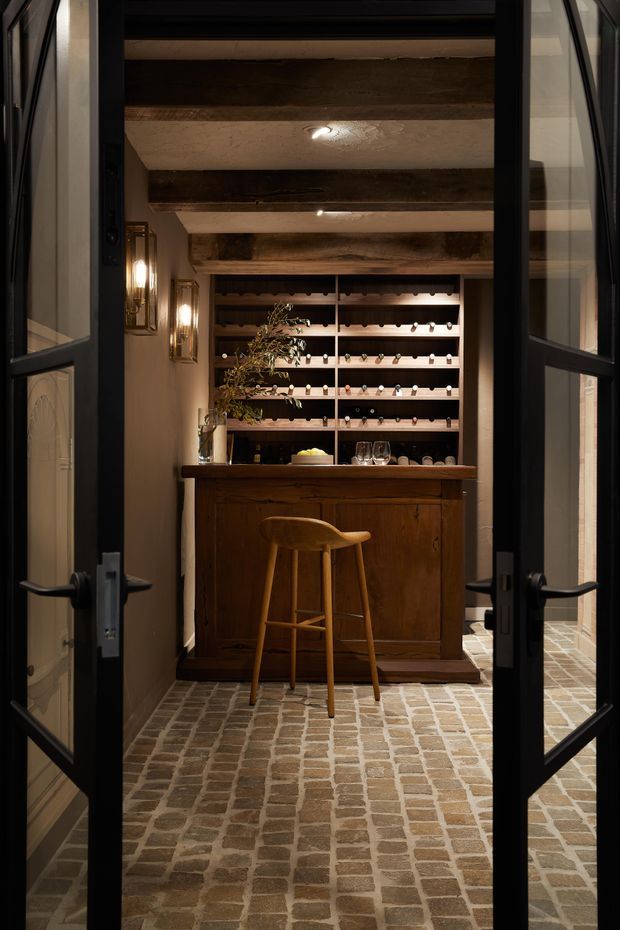 A warmly lit, compact wine room viewed through open black-framed glazed double doors. The space features exposed dark timber ceiling beams, a full-height timber wine storage wall with horizontal bottle racks, a rustic timber bar cabinet and counter, a single wooden bar stool, glassware, decorative wall sconces, and a small-format brick or stone-paver floor. The overall atmosphere is intimate, rustic, and refined.