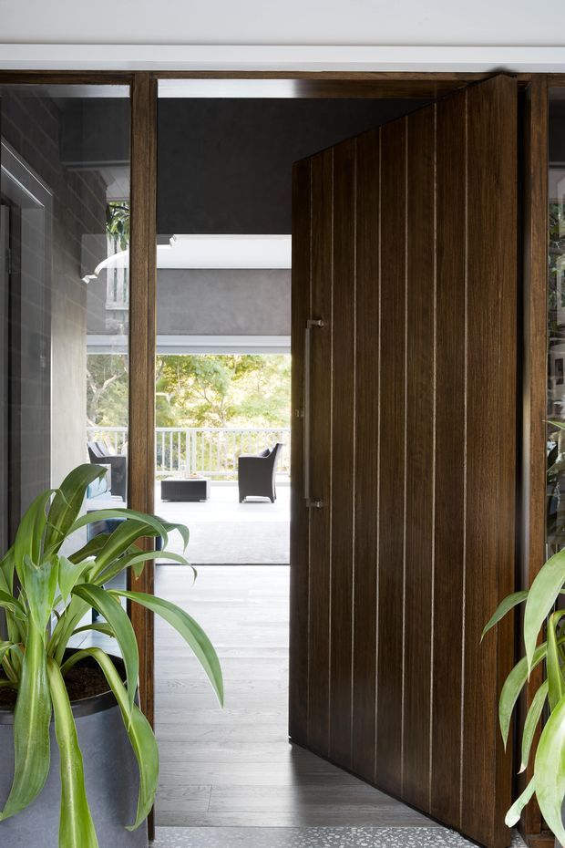 View through an entryway with a large dark timber pivot door standing open toward a bright living area and balcony. The entry has dark timber-framed glazing, grey walls, pale timber flooring, and a terrazzo-like threshold. Large leafy potted plants frame both sides, while outdoor seating and greenery are visible beyond the opening.