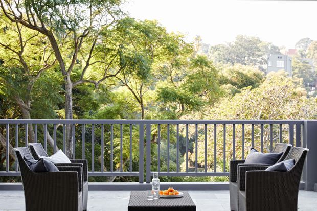 A furnished outdoor terrace or balcony with two dark woven armchairs, cushions, and a small coffee table, enclosed by a grey metal balustrade. The space overlooks a lush, mature treed landscape with a distant residential building visible beyond.