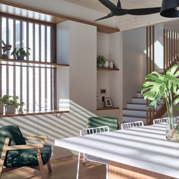 Bright, contemporary dining area with a large white dining table and white metal chairs, accompanied by a dark green timber-framed armchair. Full-height timber-framed windows with vertical screening cast strong linear shadows across the room. Built-in white cabinetry and open timber shelves provide storage and display, while timber ceiling lining, wood flooring, a sculptural black ceiling fan, and a slatted timber stair divider add warmth and texture. Indoor plants and soft daylight create a calm, airy atmosphere.
