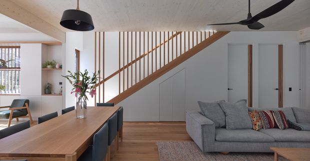A bright open-plan living and dining area with a large timber dining table and dark upholstered chairs in the foreground, a grey sectional sofa to the right, and a timber staircase with vertical slatted balustrade rising along the rear wall. White built-in cabinetry sits beneath the stairs, while pale timber ceiling boards, timber flooring, pendant lighting, and a ceiling fan create a warm contemporary interior.