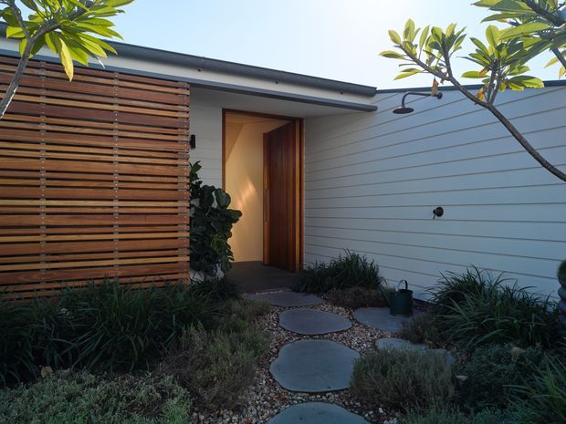 A landscaped entry approach to a low-slung modern residence, with a recessed timber front door beneath a flat roof. Horizontal timber screening sits beside pale horizontal wall cladding, while large irregular stepping stones set in gravel lead through dense architectural planting. The scene has a restrained, natural contemporary character with warm light at the entry.