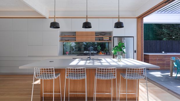 Bright contemporary kitchen with a large white island and four wire-frame bar stools, integrated pale cabinetry, timber island detailing, open timber shelving, pendant lights, and a wide opening to an outdoor entertaining area.
