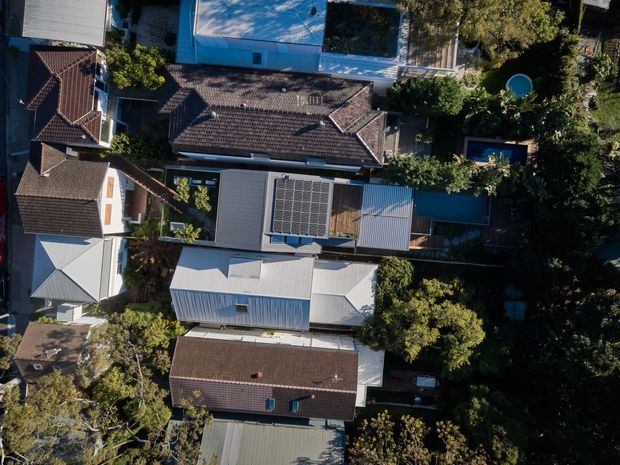 Aerial top-down view of a dense suburban residential neighbourhood, showing several detached houses with varied pitched and hipped roofs, corrugated metal roofing, tiled roofing, mature tree cover, private backyards, and multiple visible swimming pools. Solar panels are installed on the roof of a central contemporary-looking dwelling.