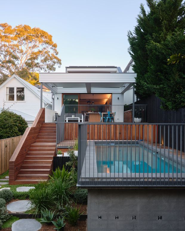 Exterior view of a contemporary multi-level home extension with a rooftop-like covered outdoor terrace, timber stairway, landscaped garden, and elevated swimming pool in the foreground. The house features crisp white cladding, dark metal-framed glazing and balustrades, a flat roof, exposed structural beams, and warm timber screening. A neighbouring or existing white weatherboard dwelling is visible to the left.