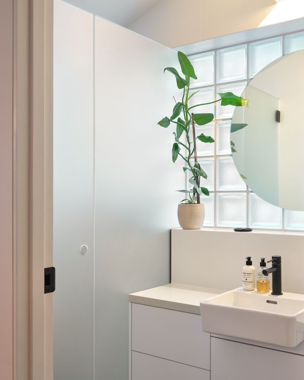 Bright, minimalist bathroom with a white wall-mounted vanity, integrated countertop, deep rectangular basin, matte black tapware, large round mirror, and a tall potted plant. A frosted glass-block and louvred window brings in abundant natural light, while pale neutral finishes create a clean, airy atmosphere.