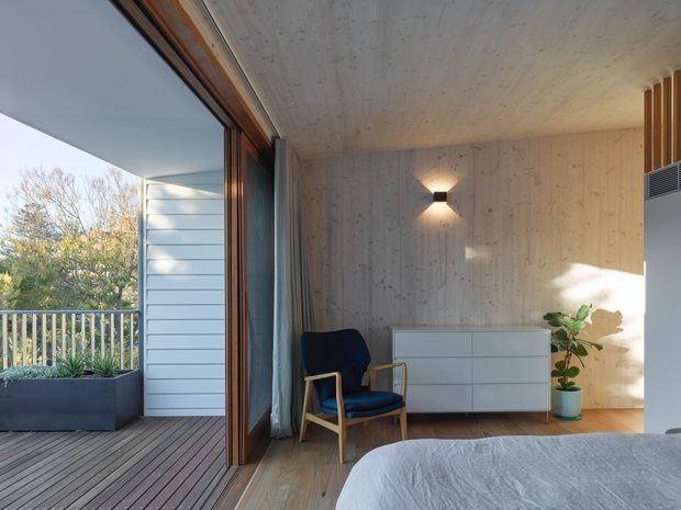 A bright, modern bedroom opening onto a timber deck through large sliding glazed doors. The room features a light timber-lined ceiling and walls, timber flooring, a low white built-in dresser, a dark upholstered wooden armchair, a potted plant, and a minimalist wall sconce. The deck has a large planter and railing overlooking leafy surroundings.
