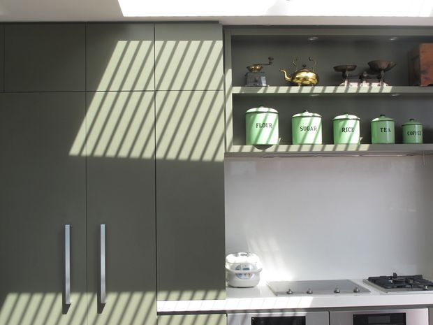 A compact contemporary kitchen with tall dark grey-green cabinetry, long vertical brushed-metal handles, open display shelving, and a pale smooth backsplash. The shelves hold labelled green storage canisters and decorative vessels, while a cooktop and built-in oven are visible below. Strong diagonal sunlight and shadows from overhead glazing create a high-contrast graphic pattern across the cabinetry and work surfaces.
