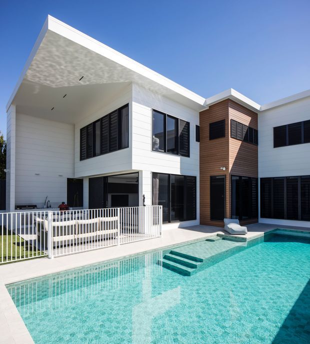 Bright exterior view of a contemporary multi-storey residence arranged around a swimming pool courtyard. The house has white horizontal cladding, dark-framed glazing with louvred screens, a warm timber-clad vertical section, projecting flat roof forms, and a covered outdoor entertaining area beside the pool.