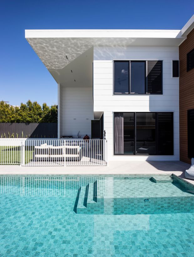 Bright contemporary two-storey residential rear exterior with white horizontal cladding, projecting flat roof and dark-framed glazing, photographed beside a turquoise tiled swimming pool. A covered outdoor patio with white balustrading and outdoor seating occupies the ground level, while a timber-clad section is visible at right.