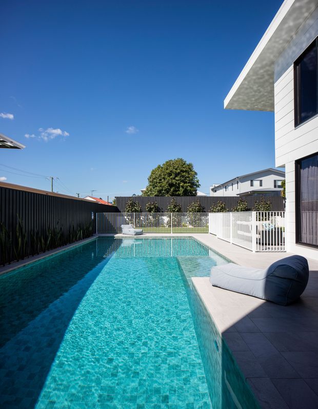A bright modern backyard pool area beside a white-clad house. The long rectangular swimming pool has turquoise mosaic tiling, a paved grey-tile terrace, a sculptural outdoor lounge seat, white metal pool fencing, and dark privacy fencing. The image is photographed under a clear blue sky.