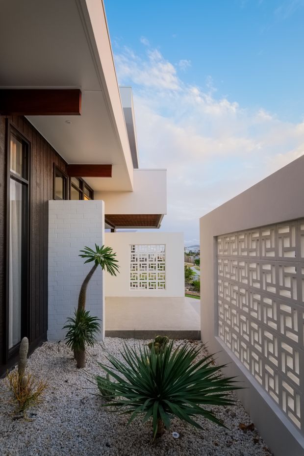 A narrow contemporary courtyard passage beside a modern multi-level residence. The space features dark vertical timber cladding, crisp white rendered walls, projecting flat-roofed balconies, timber soffit accents, and decorative white breeze-block screens. A gravel xeriscape garden with sculptural yucca-like plants runs along the walkway, opening toward a distant urban or coastal view under a blue sky.