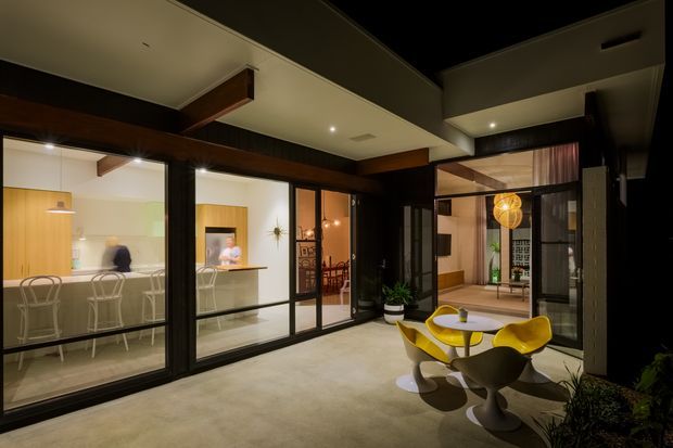 A dark, sophisticated courtyard or covered outdoor entertaining area framed by extensive black-framed glazing and timber beams. The space contains a small round table with distinctive yellow sculptural chairs, potted plants, and views into a bright kitchen, dining area, and interior living spaces. The building features a modern open-plan composition with strong indoor-outdoor connection, exposed dark timber structural elements, and a layered ceiling with recessed lighting.
